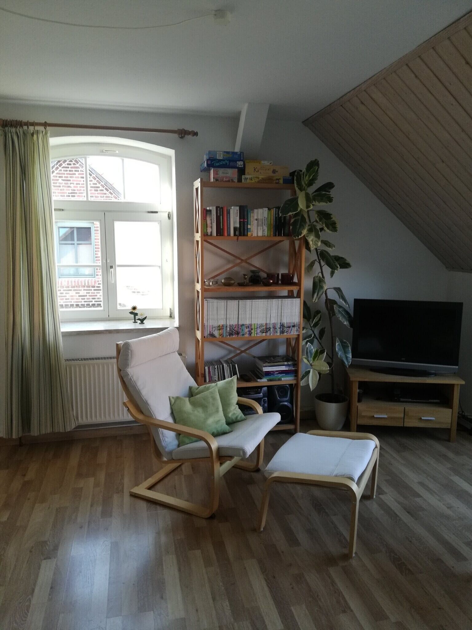 Un salon avec un plancher en bois, un fauteuil à bascule, une bibliothèque, une plante en pot, un téléviseur et une fenêtre avec des rideaux.