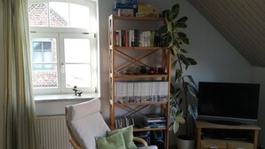 Een woonkamer met houten vloer, een schommelstoel, een boekenplank, een kamerplant, een televisie en een raam met gordijnen.