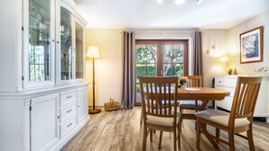 Een eetgedeelte met een houten tafel en stoelen, een wit kastje met glazen deuren, een vloerlamp en uitzicht door een raam met gordijnen.