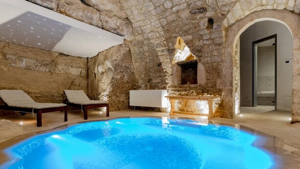 A stone-walled indoor pool with a blue-lit bottom, lounge chairs, and a modern fireplace.