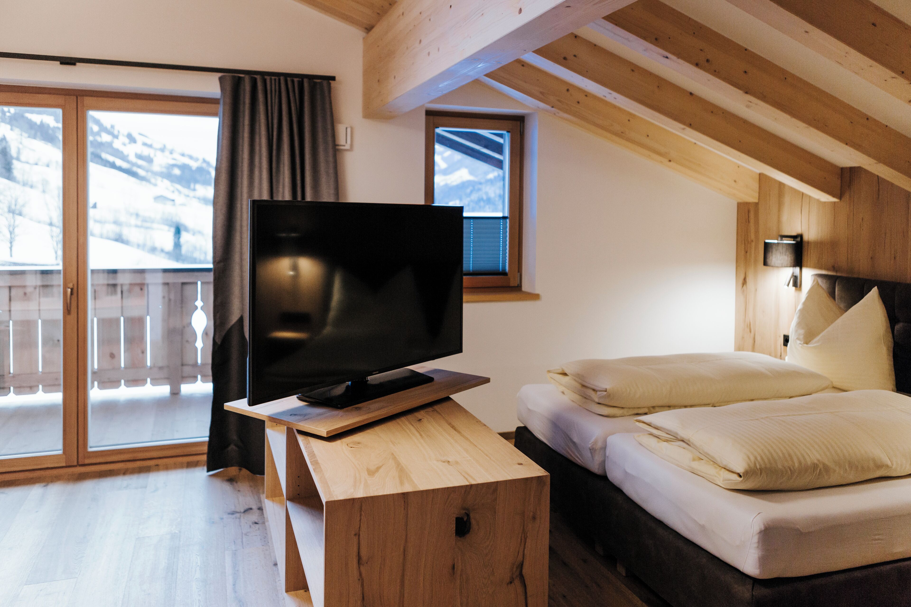 A bedroom with a wooden bed, a TV on a wooden stand, a balcony with a view of snow-covered mountains, and a wooden ceiling.