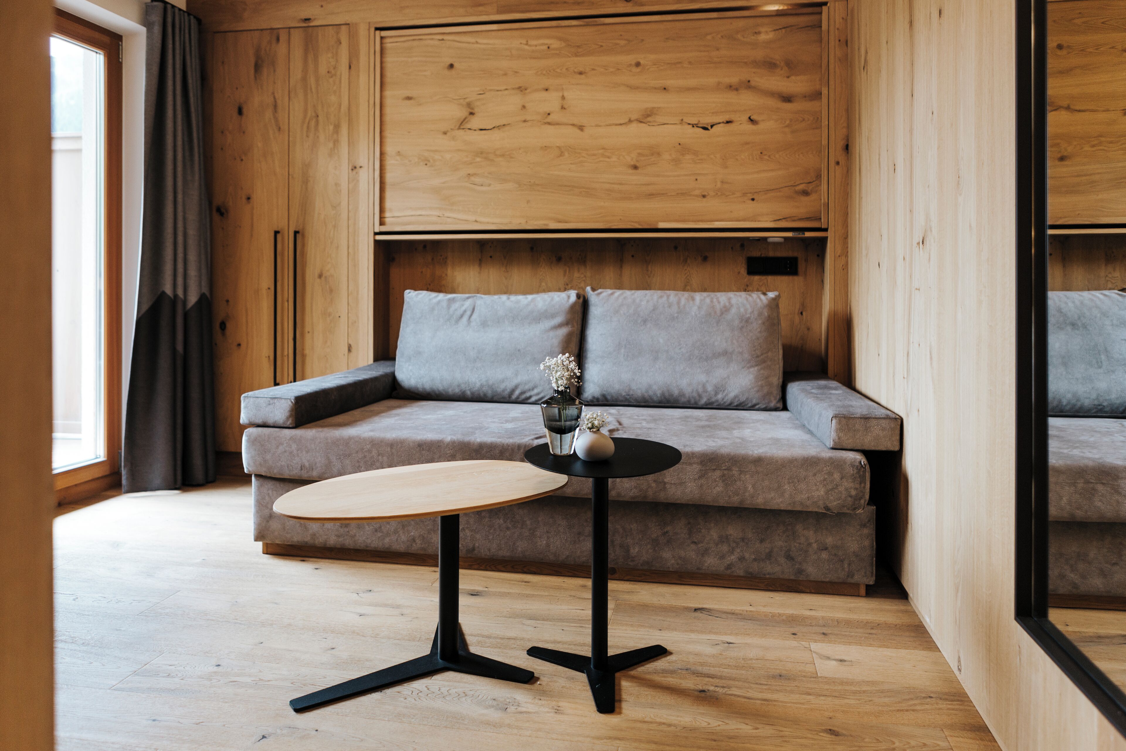 Ein modernes Zimmer mit Holzboden, einem grauen Sofa, einem kleinen runden Tisch und einem Fenster mit Vorhängen.