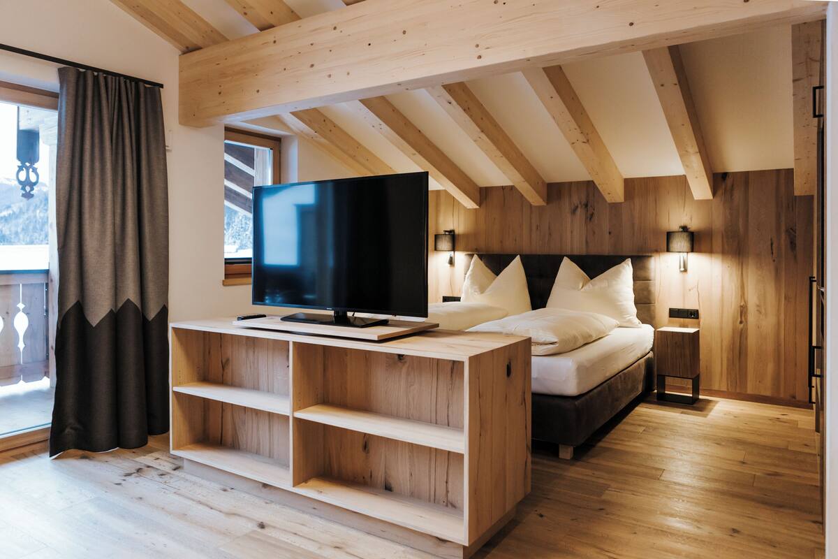 Een moderne slaapkamer met een houten plafond, een bed met wit beddengoed, een televisie op een houten dressoir en een nachtkastje met een lamp.