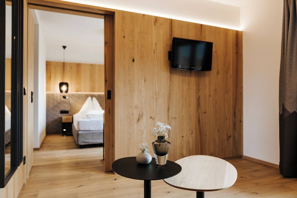 Een moderne hotelkamer met een houten vloer, een ronde tafel met een bloevas, een bed met een wit hoofdeinde, en een aan de muur bevestigde televisie.