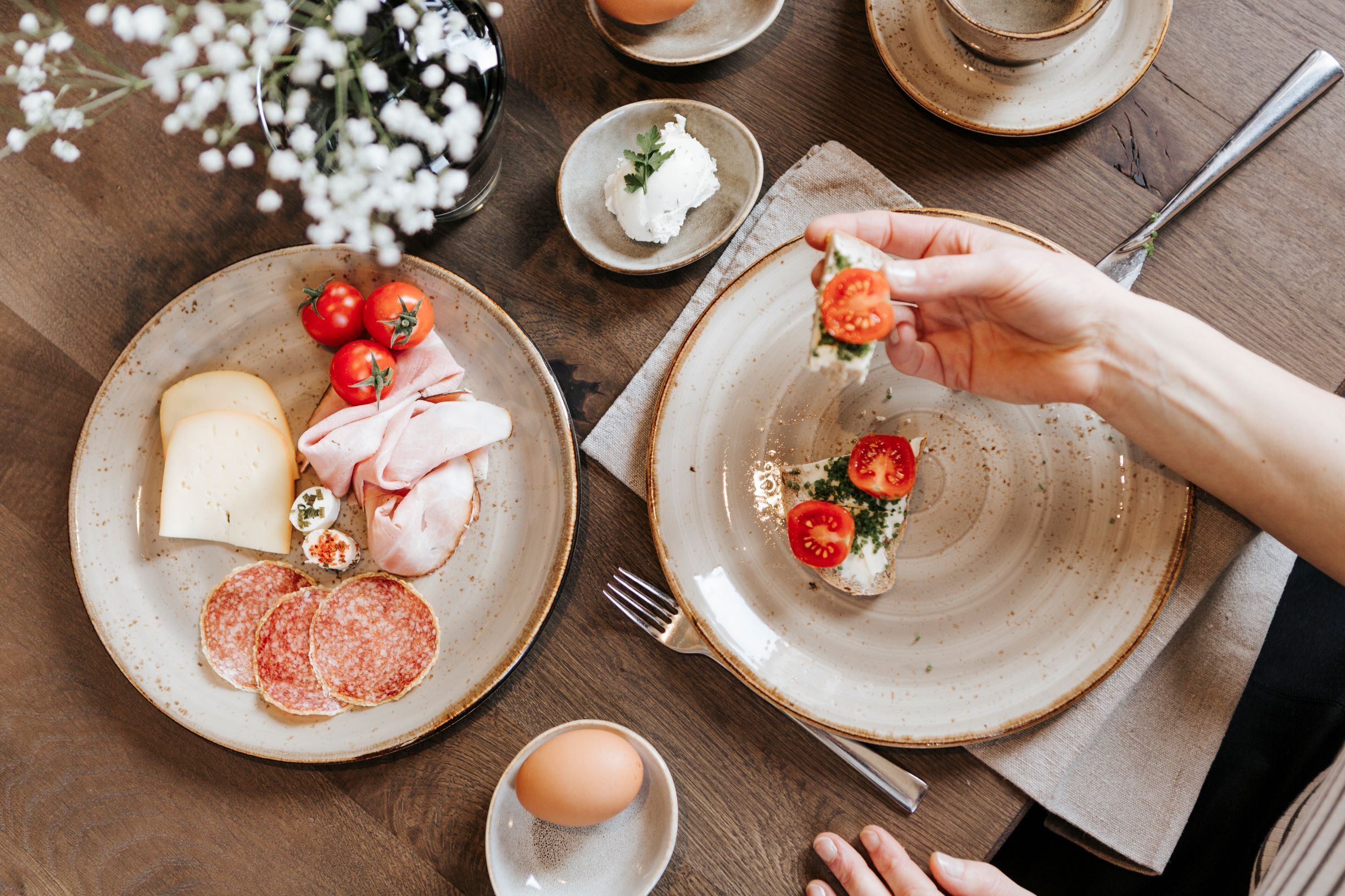 Una mesa servida con platos de queso, jamón, salami y tomates, con una mano escogiendo un tomate.