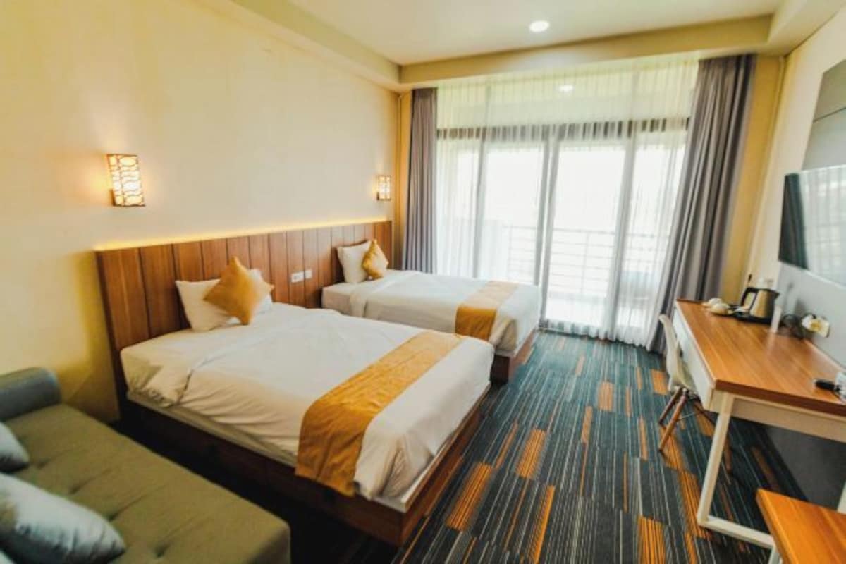 A hotel room with two beds, a desk, a TV, and a view of the outdoors.