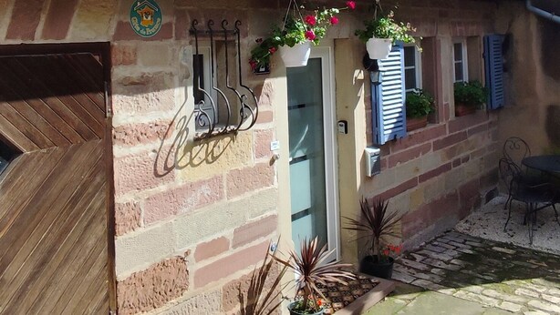 Un edificio de dos pisos con techo de tejas, fachada de piedra y puerta de madera. Hay plantas en macetas en la planta baja y un perchero metálico con objetos colgando encima de la puerta.
