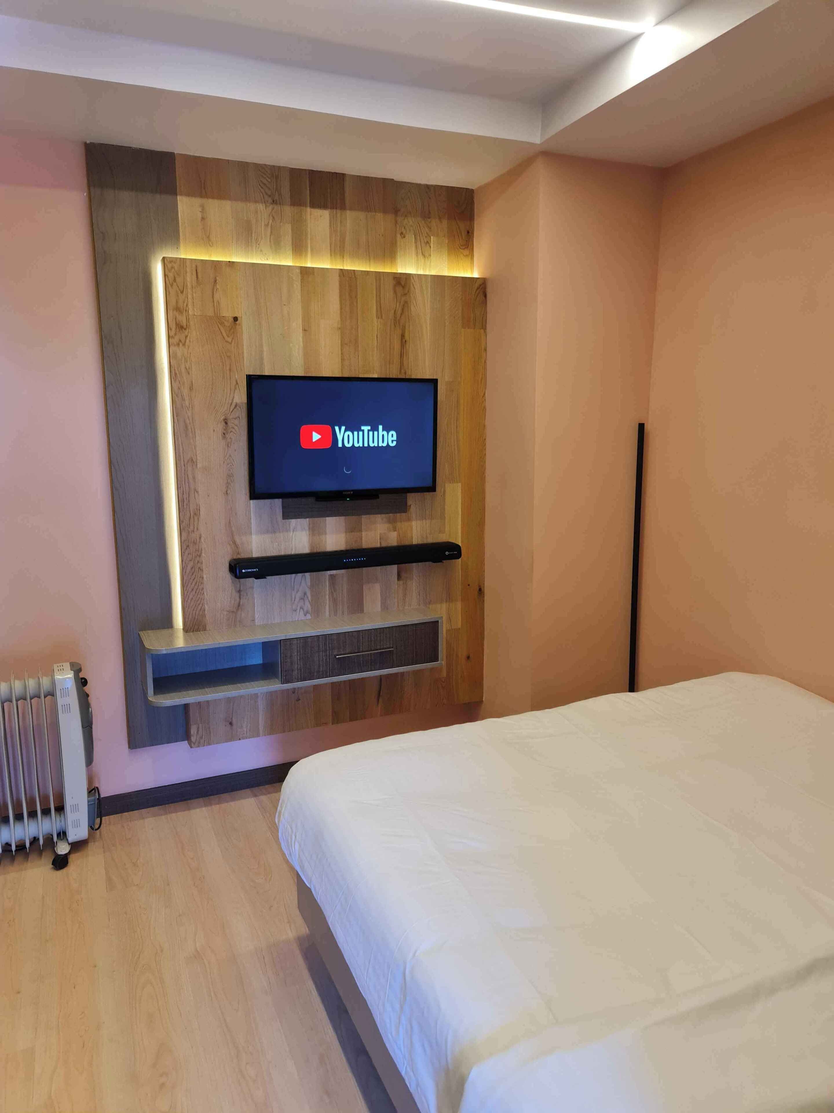 A modern hotel room with a wooden accent wall, a mounted flat-screen TV, a built-in shelf, and a bed with a white duvet.