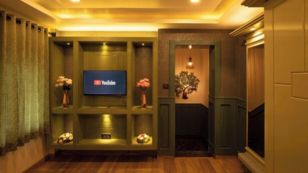A cozy living room with a flat-screen TV mounted on a green wall, wooden flooring, and a decorative tree wall art piece. - HERITAGE VIEW (Ootacamund)