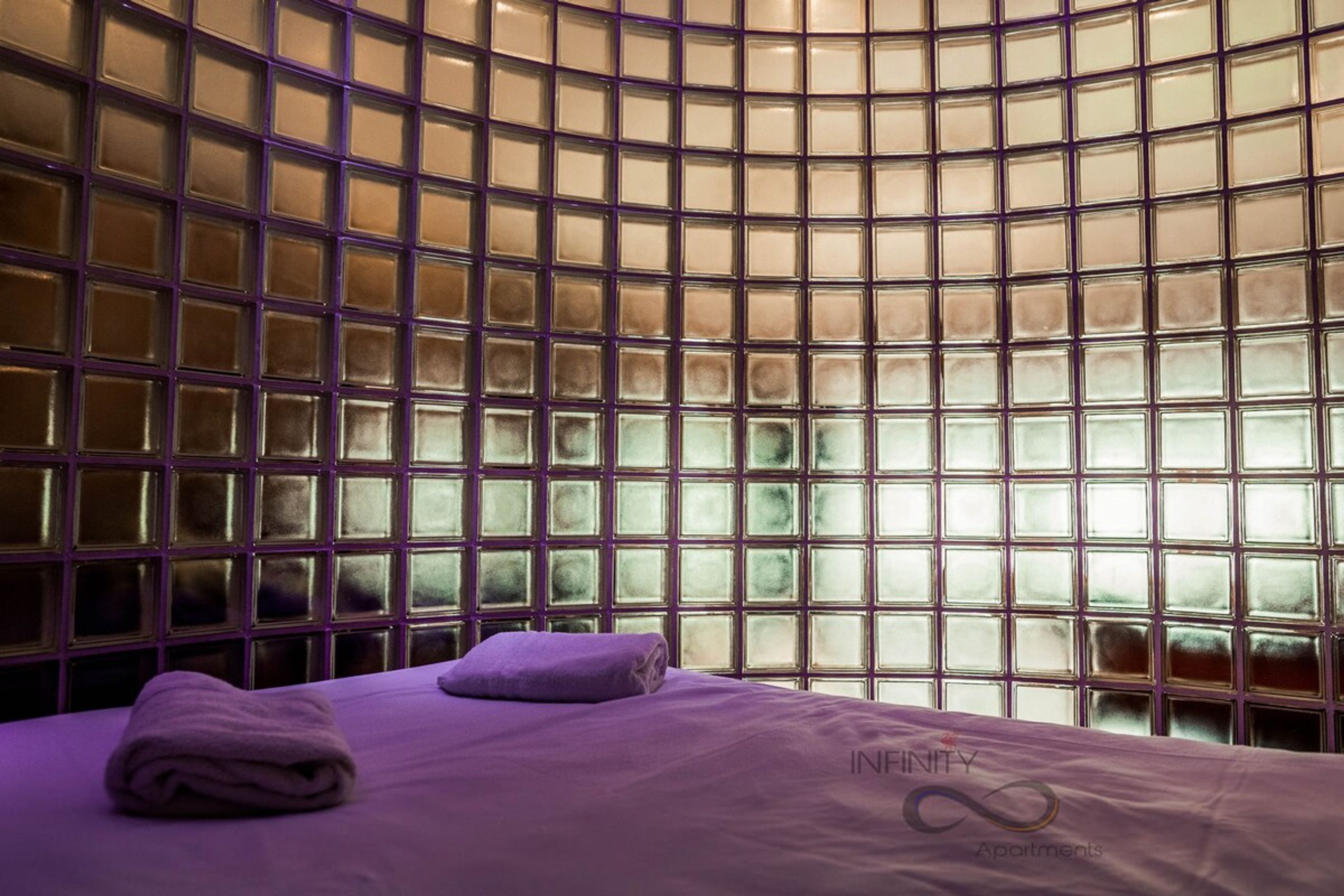 A room with a curved glass partition, a bed with a purple bedspread, and a towel on the bed.