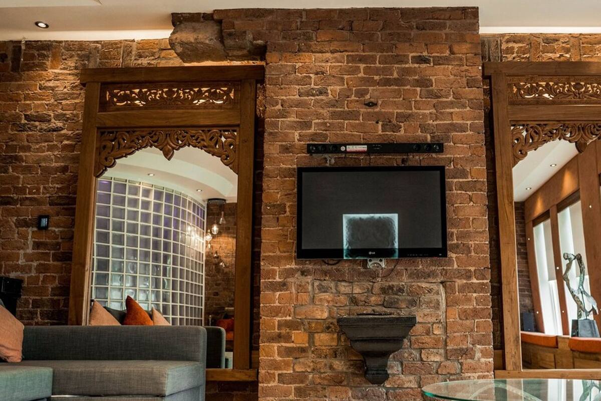 A living room with a brick wall, a flat-screen TV mounted on the wall, a glass coffee table, and a sofa with cushions.