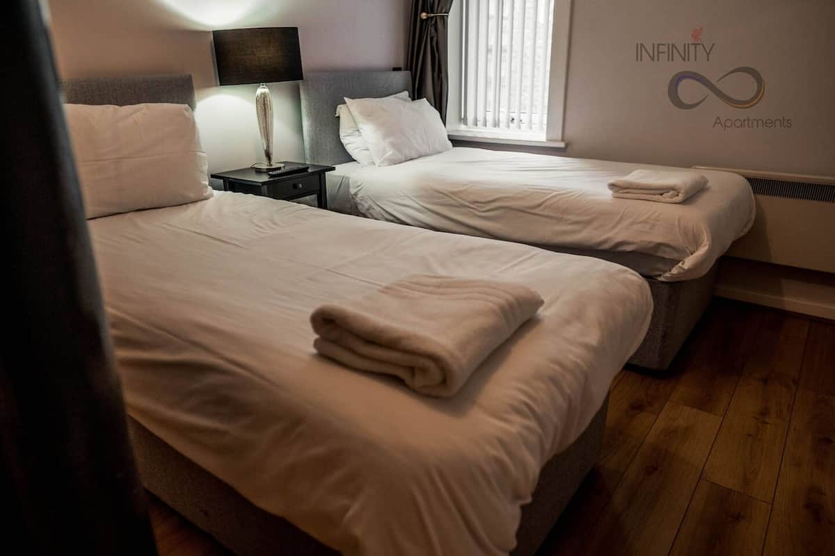 A hotel room with two beds, a nightstand, a lamp, and a window with blinds.
