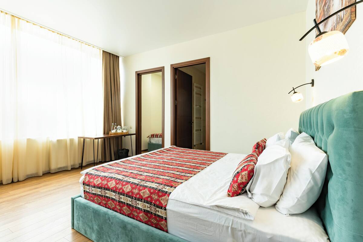 Une chambre d’hôtel avec un lit, un bureau et une chaise. Il y a une fenêtre avec des rideaux et une porte à l’arrière-plan.