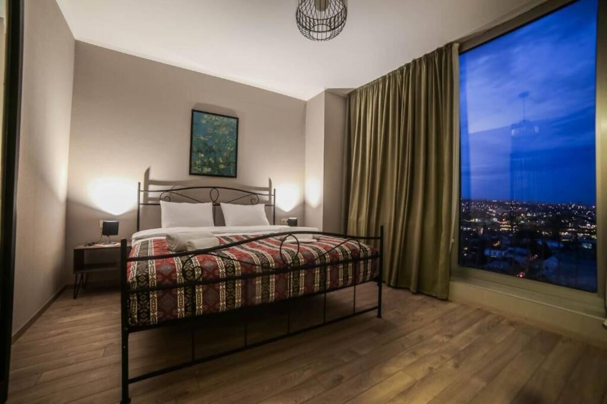 Une chambre à coucher avec un lit en métal, une table de chevet, une lampe de chevet, un tableau au mur et une grande fenêtre donnant sur la ville.