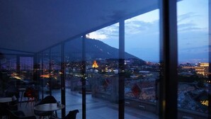 Une chambre située dans un immeuble de grande hauteur, dotée de grandes fenêtres donnant sur une vue panoramique de la ville au crépuscule.