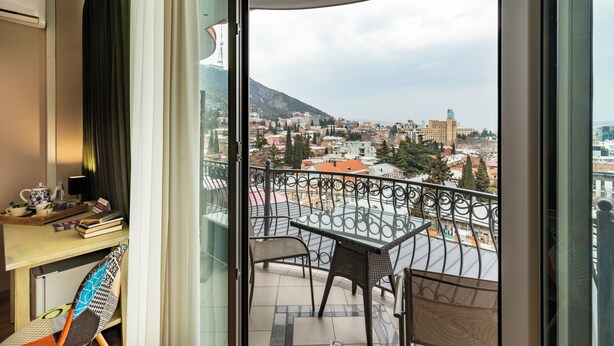 Un balcon avec une table et des chaises, un bureau avec des livres et une théière, et une vue sur un paysage urbain avec des bâtiments et une colline.