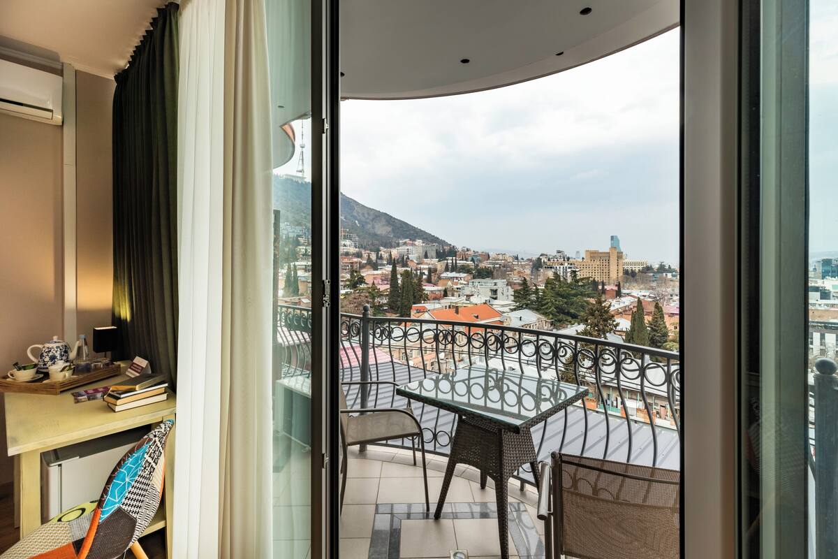 Un balcon avec une table et des chaises, un bureau avec des livres et une théière, et une vue sur un paysage urbain avec des bâtiments et une colline.