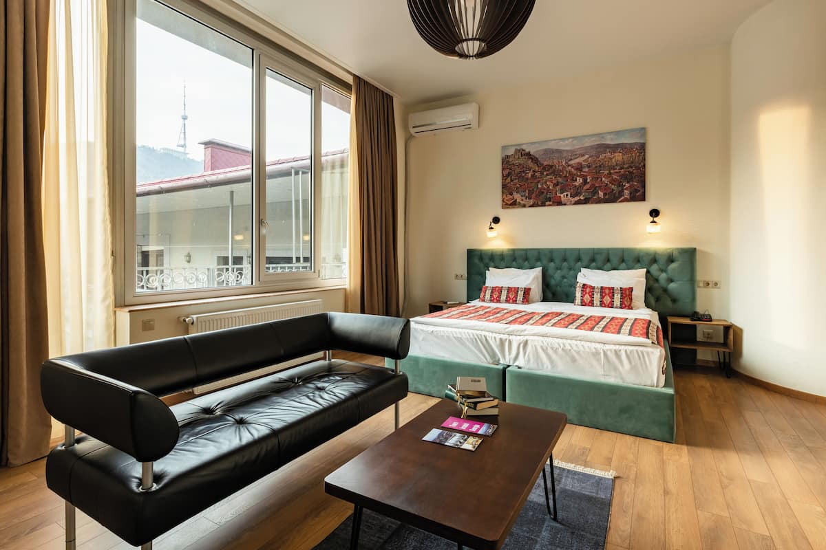 Une chambre d’hôtel moderne avec un lit aux couleurs vertes, un canapé en cuir noir, une table basse en bois et un tableau représentant un paysage urbain accroché au mur.