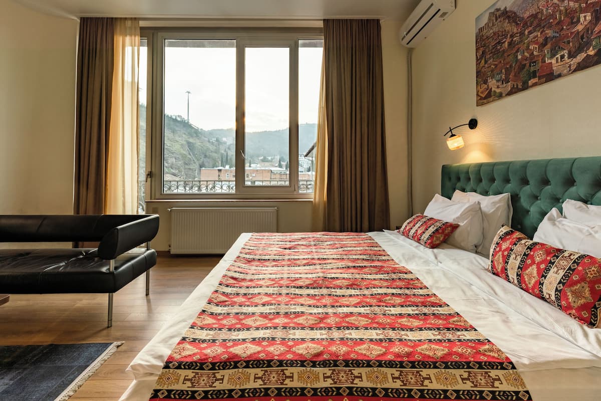 Une chambre à coucher avec un grand lit, une tête de lit recouverte d’un tissu vert et une vue sur une colline, visible par la fenêtre.
