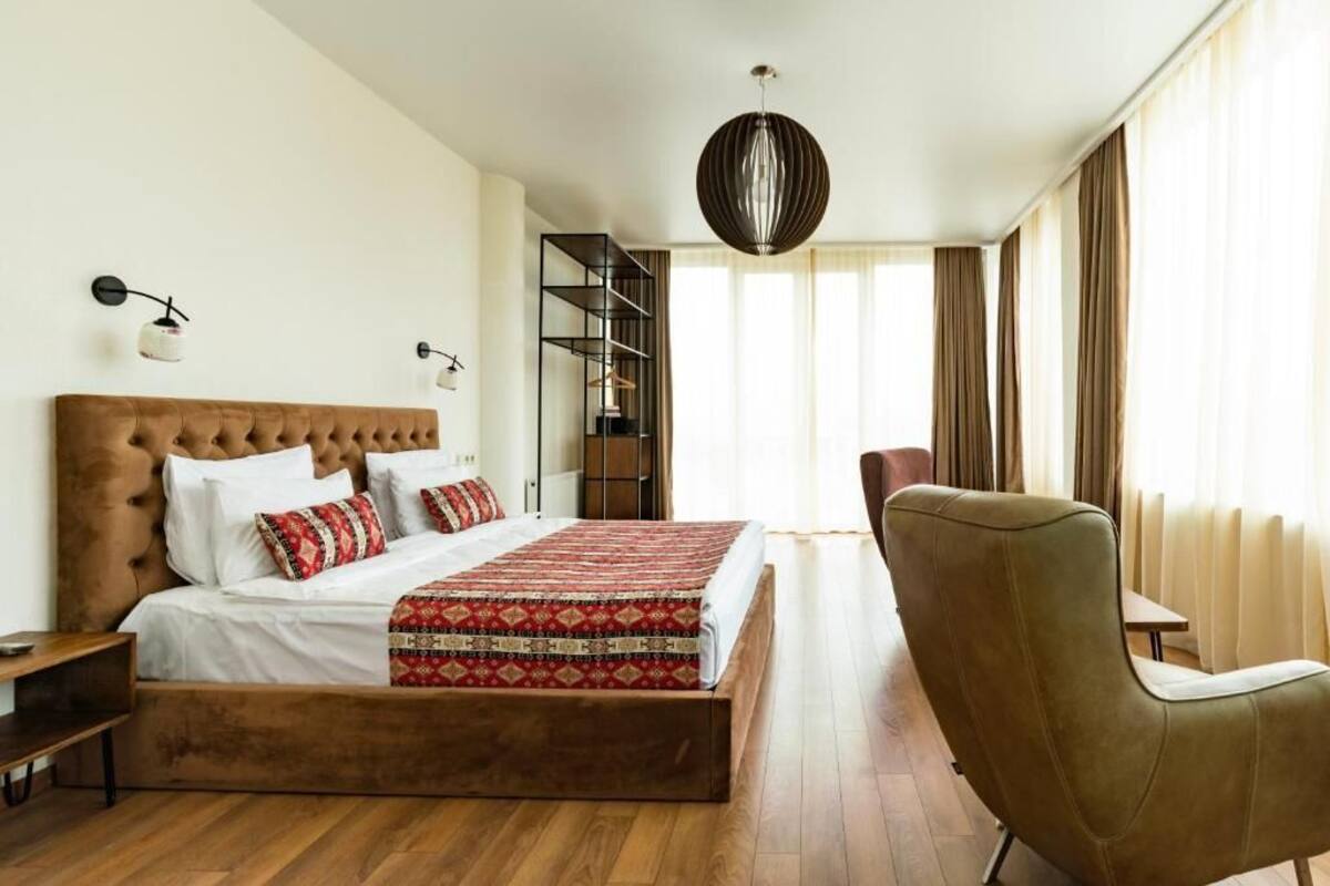Une chambre à coucher avec une tête de lit capitonnée, un grand lit avec une literie à motifs, une table de chevet, une chaise, un luminaire suspendu et une vue sur un balcon avec des rideaux.
