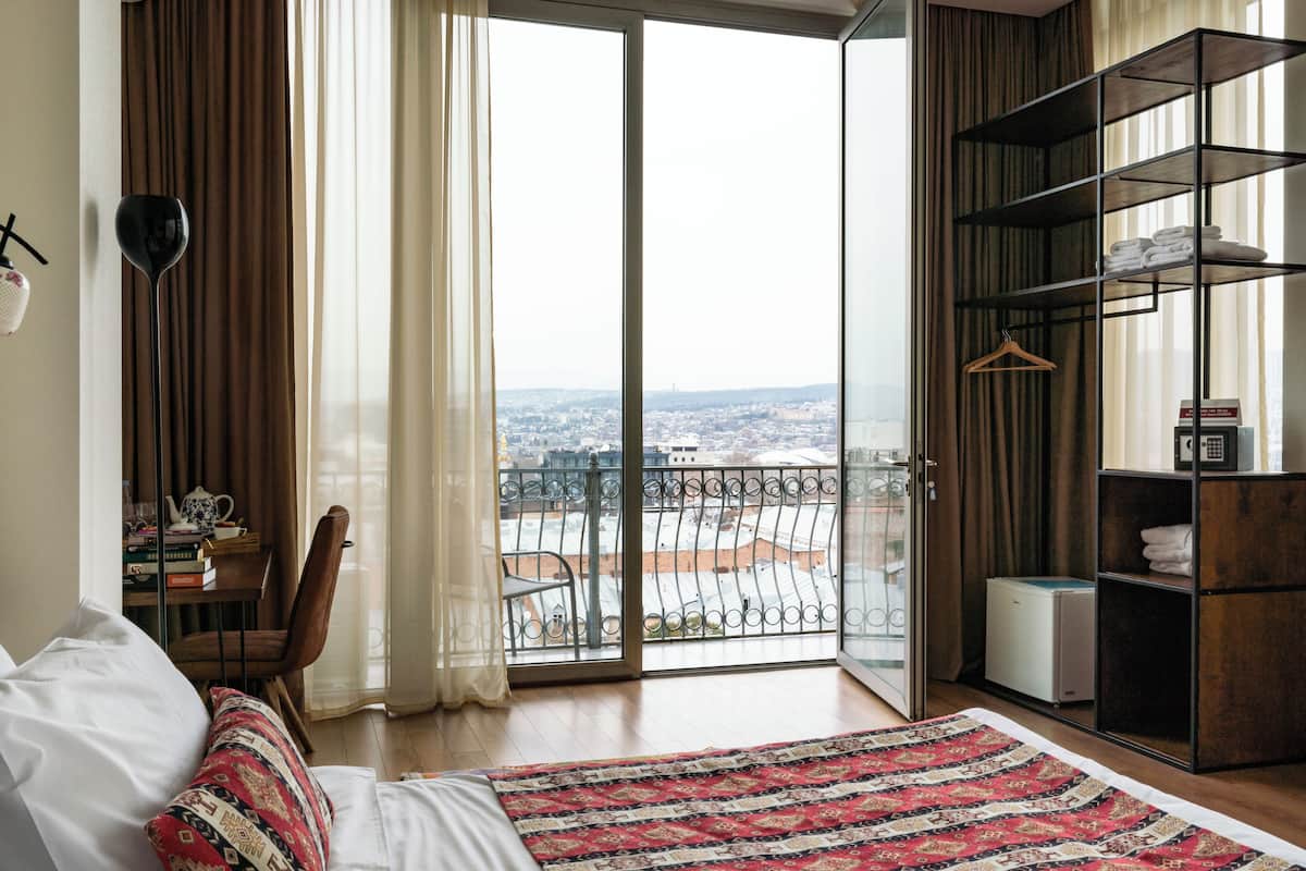 Une chambre avec un grand lit, une vue sur la ville grâce à une porte-fenêtre coulissante, une bibliothèque et un fauteuil.