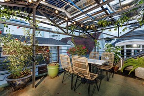 Ulkoterassi, jossa on pöytä ja tuoleja, ja jota ympäröivät ruukuissa olevat kasvit sekä köynnöskasveilla koristeltu pergola, johon on ripustettu valonauhoja.