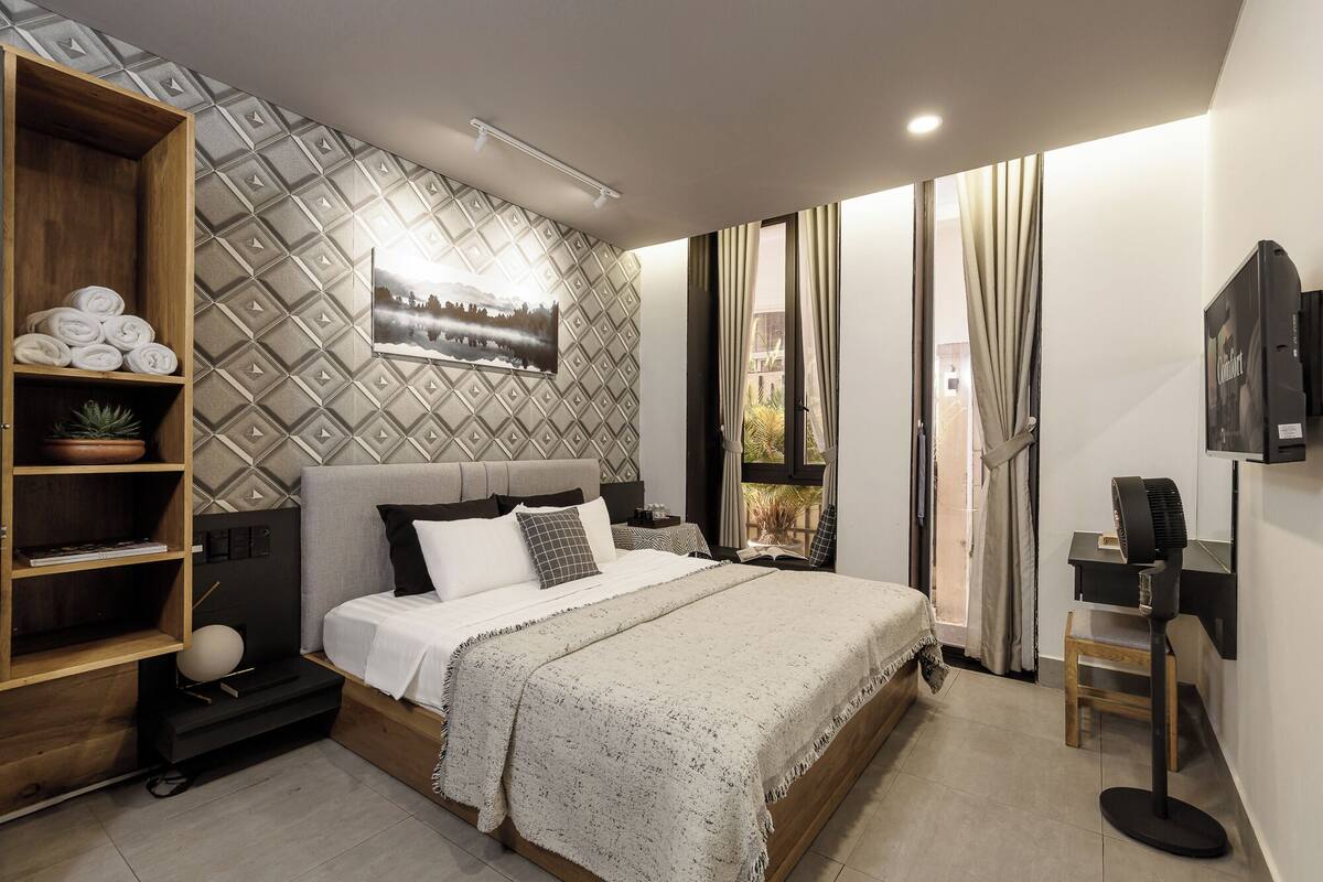 A modern bedroom with a bed, a wooden shelf with towels, a potted plant, a wall-mounted TV, and a window with curtains.