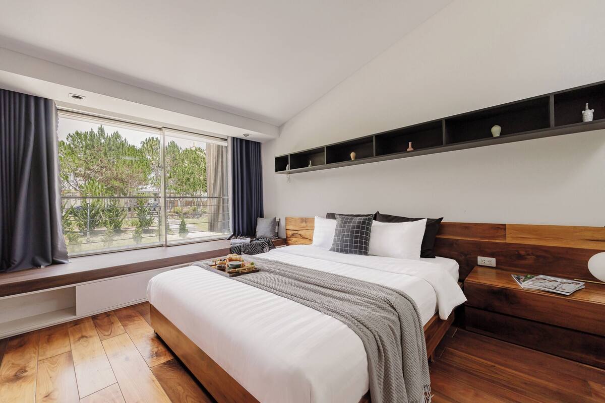 A modern bedroom with a large window, a bed, a nightstand, and a wooden headboard.