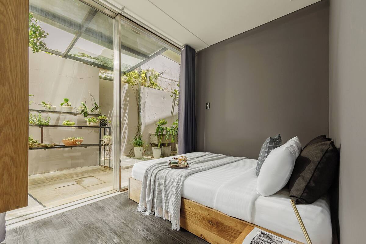 A bedroom with a bed, a bedside table, a window, and a view of a balcony with plants.
