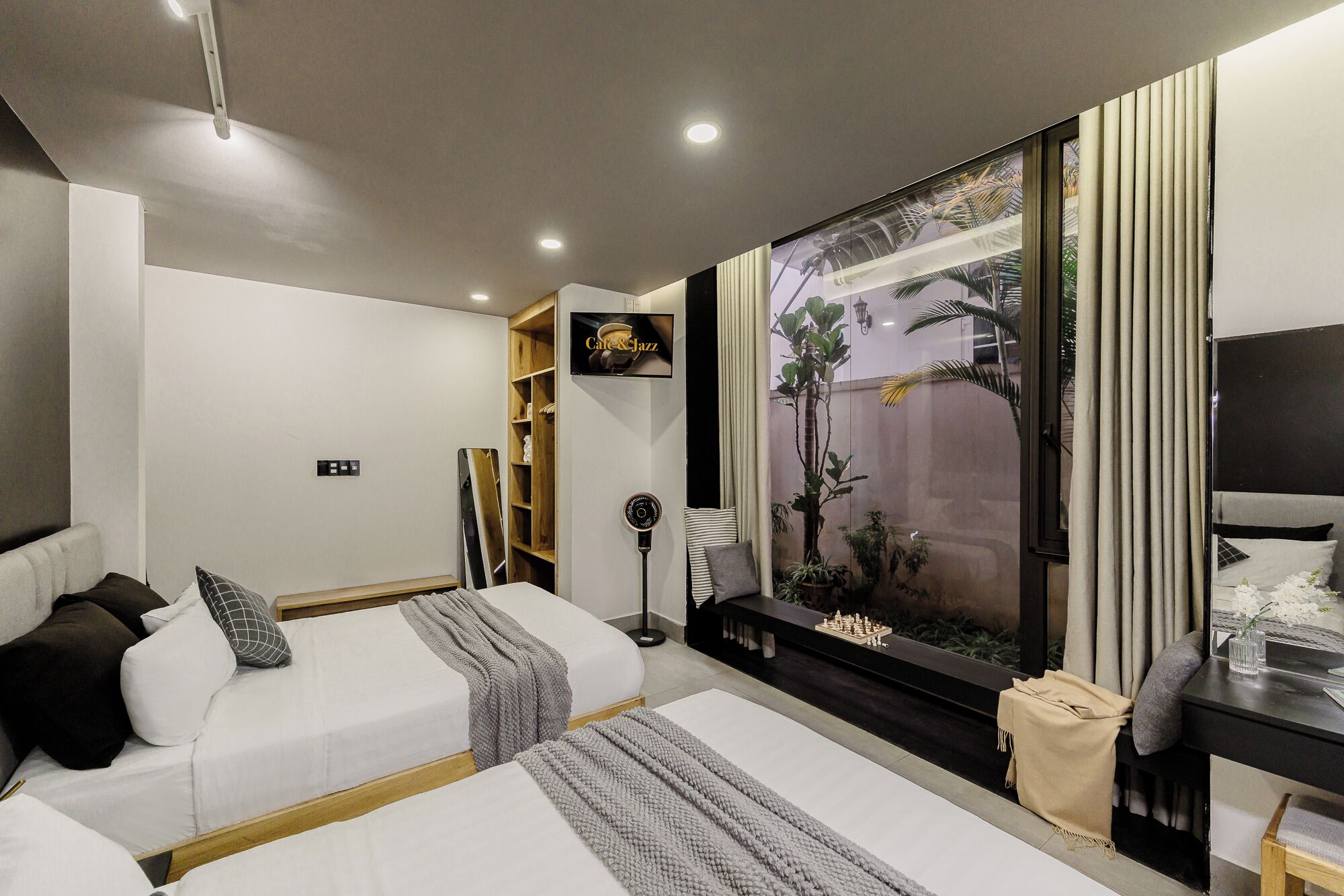 Une chambre d’hôtel moderne équipée de deux lits, d’une télévision, d’une grande fenêtre donnant sur des palmiers et d’un petit bureau avec une lampe.