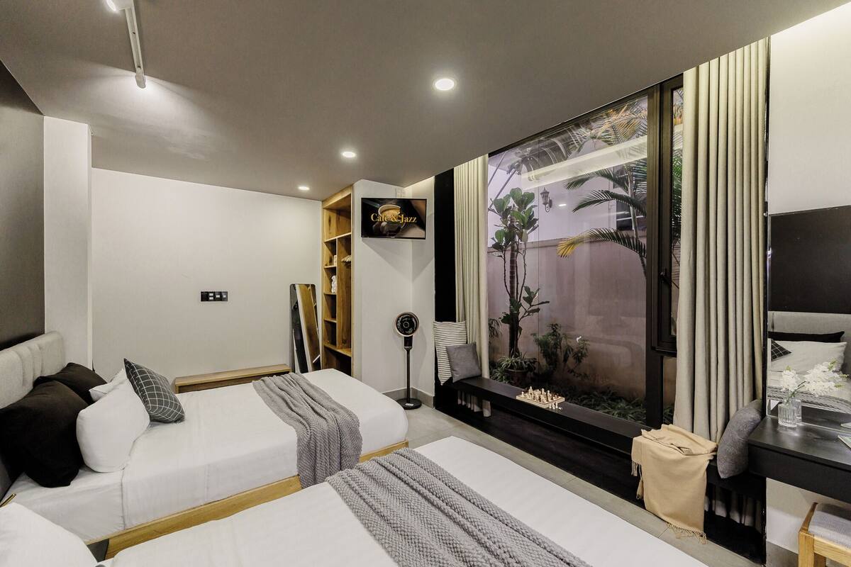A modern hotel room with two beds, a TV, a large window with a view of palm trees, and a small desk with a lamp.