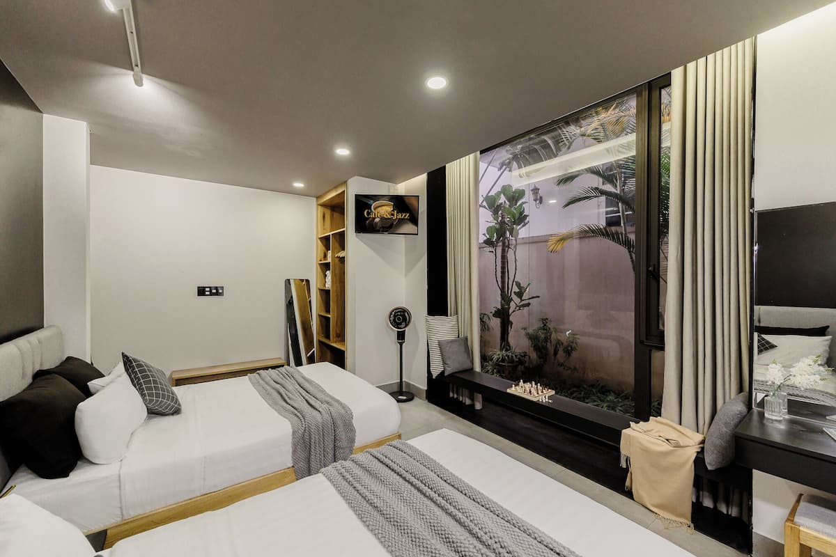 Una habitación de hotel moderna con dos camas, un televisor, un ventanal con vistas a palmeras y un pequeño escritorio con lámpara.