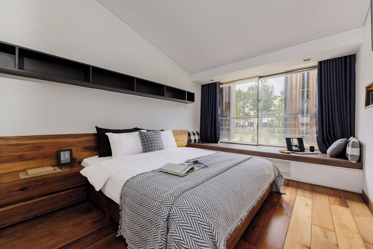 A modern bedroom with a wooden bed, a window seat with a laptop, and a view of buildings outside.
