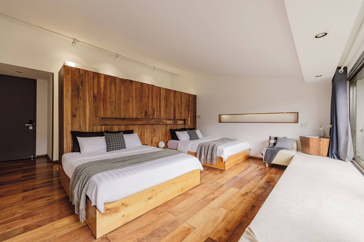 A modern bedroom with two beds, wooden flooring, a large wooden headboard, and a minimalist decor.