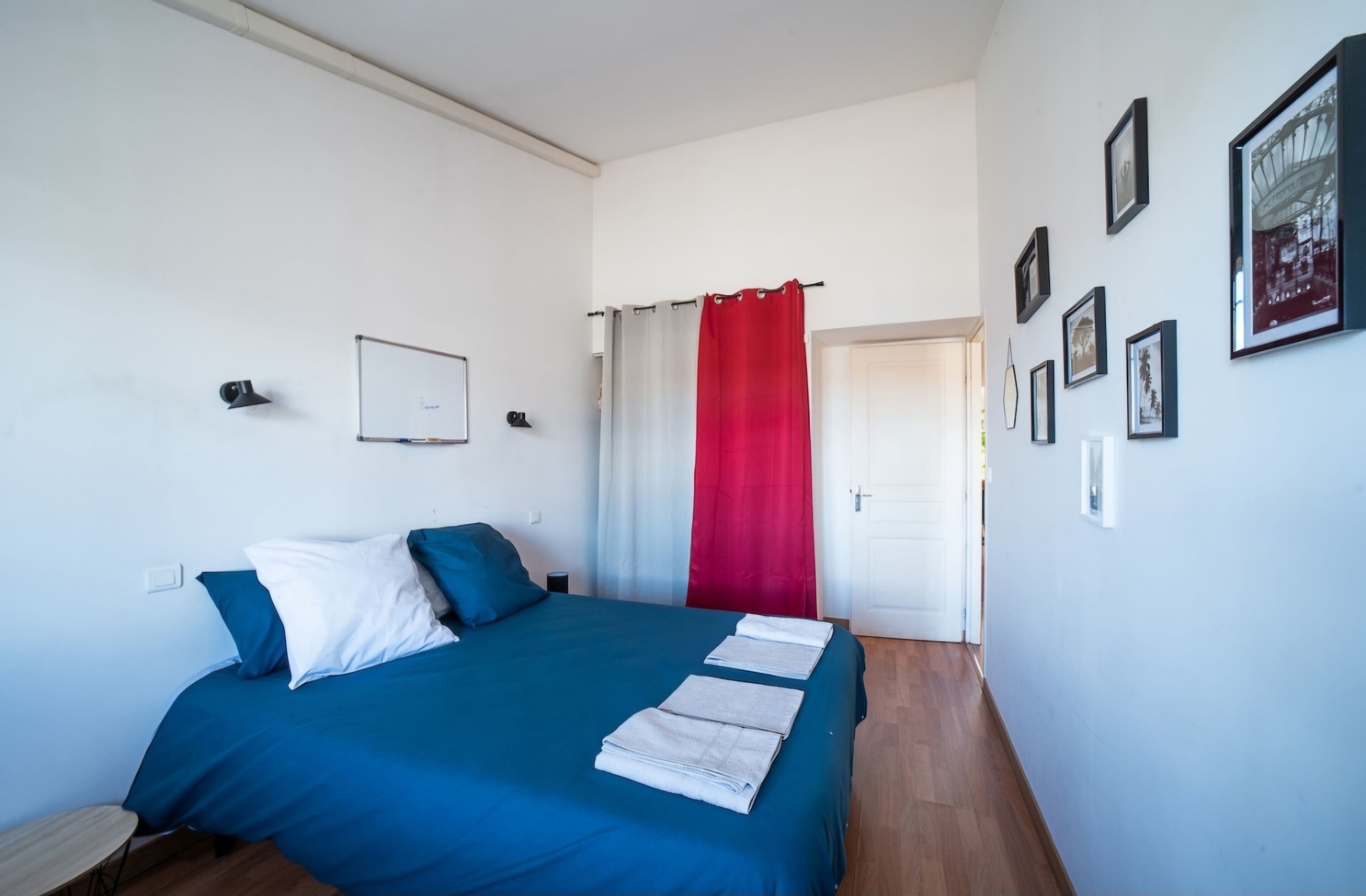 Une chambre à coucher avec un lit, une table de chevet, un rideau rouge et des tableaux encadrés accrochés au mur.