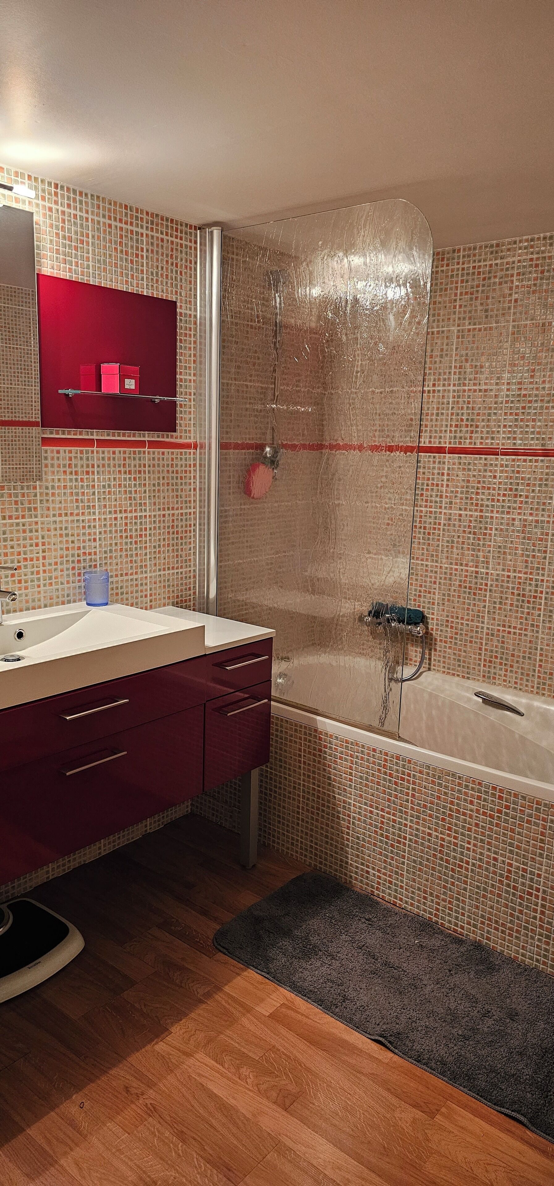 A bathroom with a wooden floor, a white sink, a red cabinet, a glass shower enclosure, and a tiled wall.