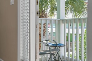 A balcony with a metal chair and a small table, a white railing, and a view of a green building and palm trees.