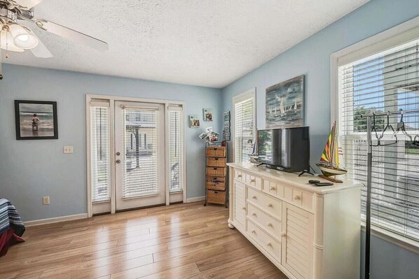 A room with a wooden floor, a ceiling fan, a TV on a white cabinet, and a window with blinds.
