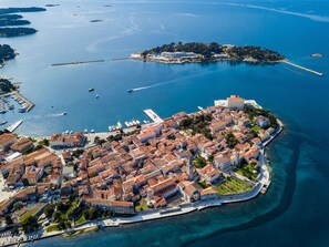 Vue aérienne d’une ville côtière avec un ensemble dense de bâtiments, un imposant bâtiment blanc surmonté d’un toit rouge, et une marina où sont amarrés des bateaux.