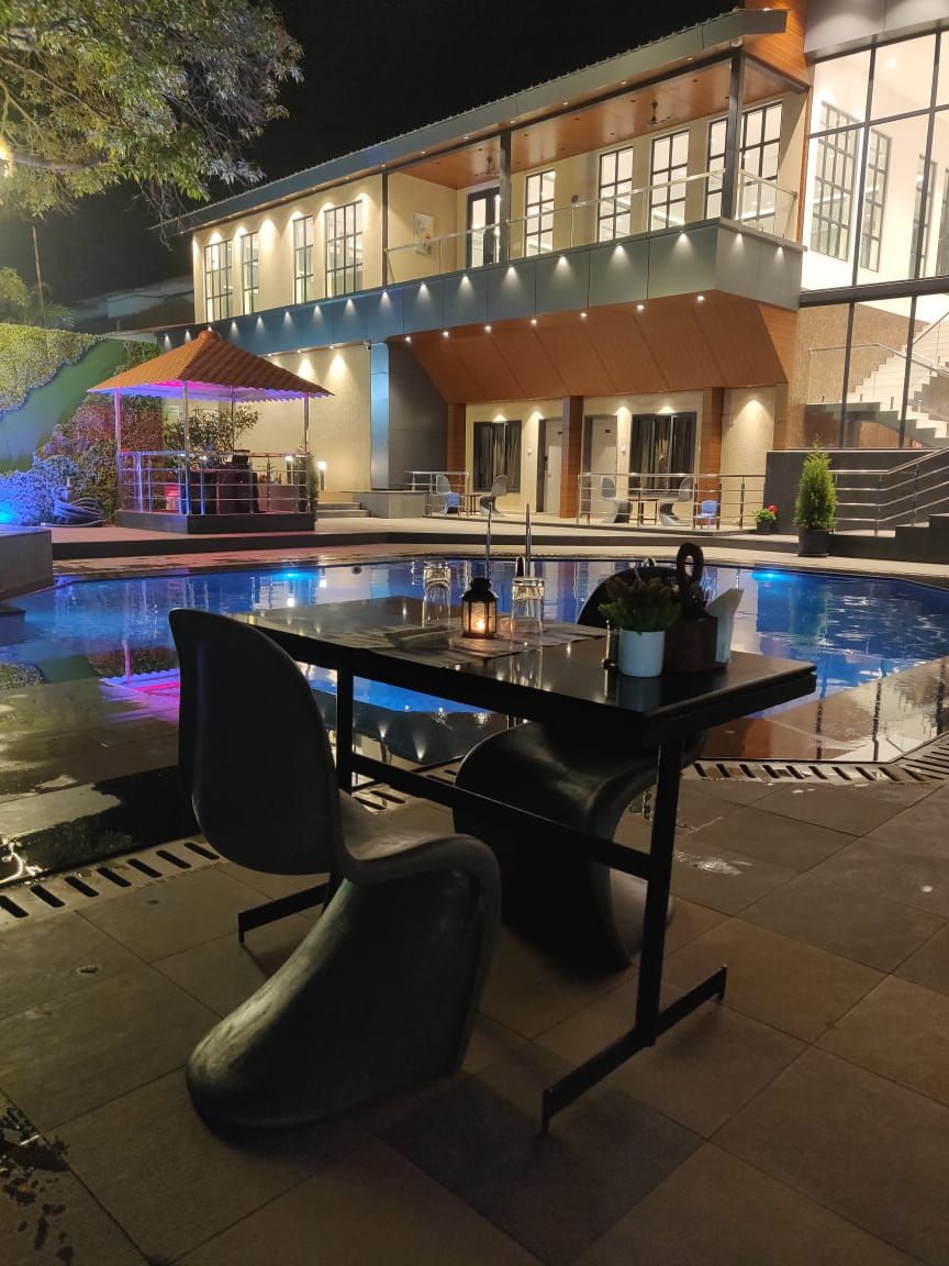 Et moderne poolområde med spisebord, stole, en pool og en bygning med flere vinduer samt et overdækket siddeområde.