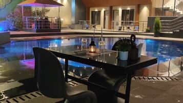 Et moderne poolområde med spisebord, stole, en pool og en bygning med flere vinduer samt et overdækket siddeområde.