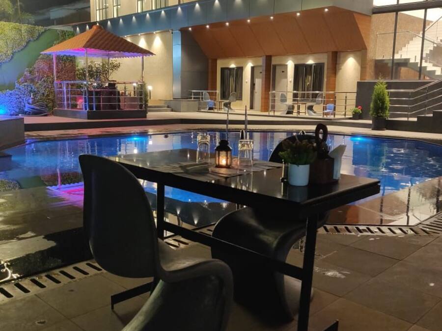 Et moderne poolområde med spisebord, stole, en pool og en bygning med flere vinduer samt et overdækket siddeområde.