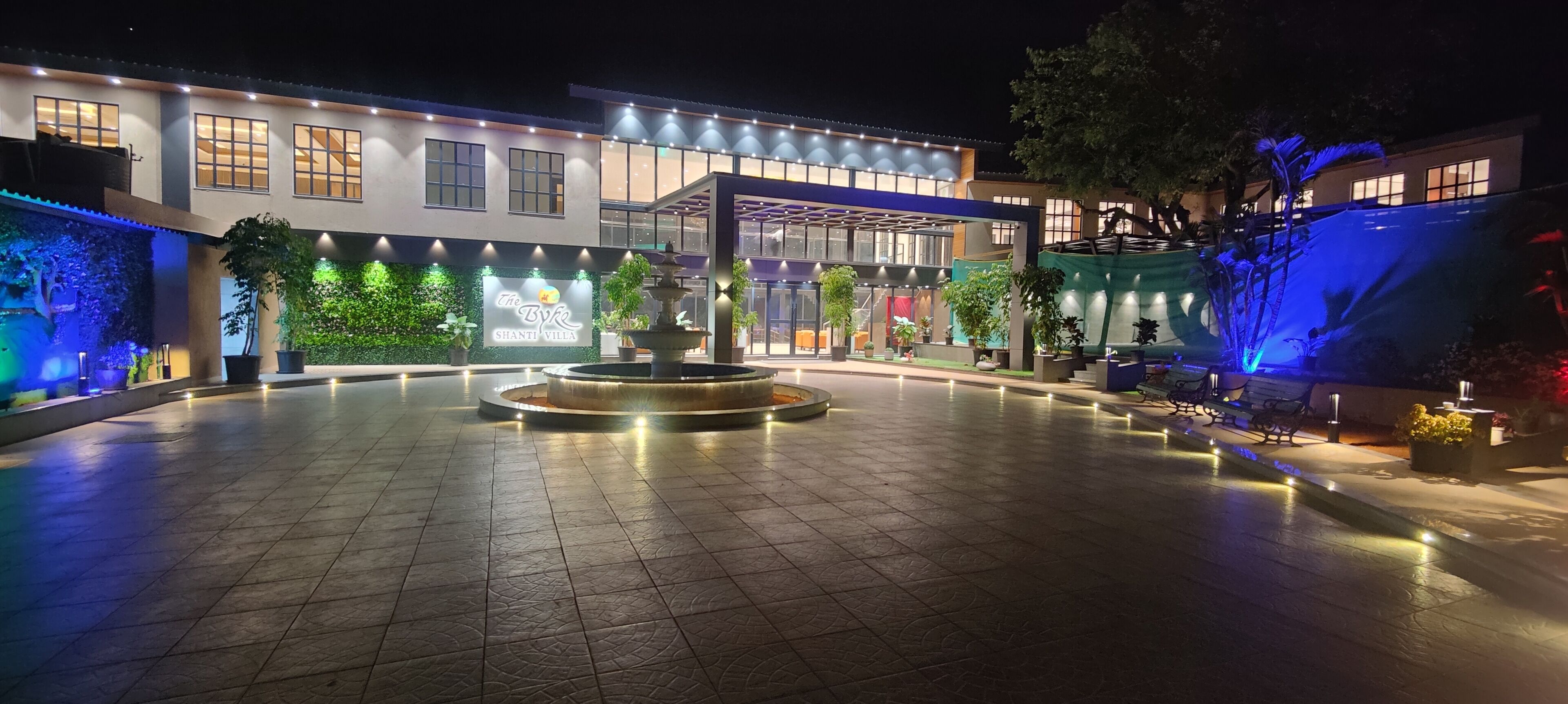 A well-lit courtyard with a central fountain, surrounded by potted plants and a modern building facade.