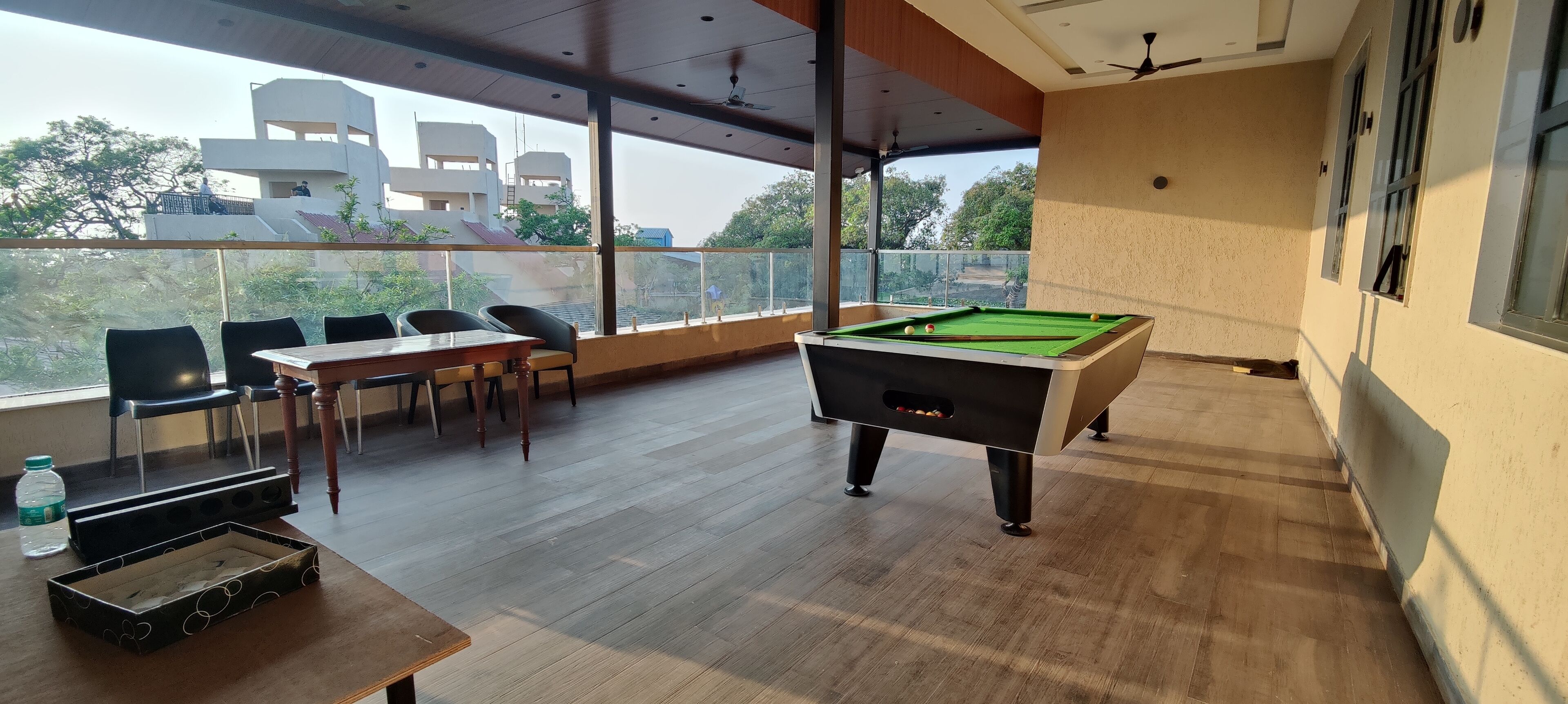 A pool table in a room with a view of buildings and trees. There are chairs and a table with a bottle and a tray.