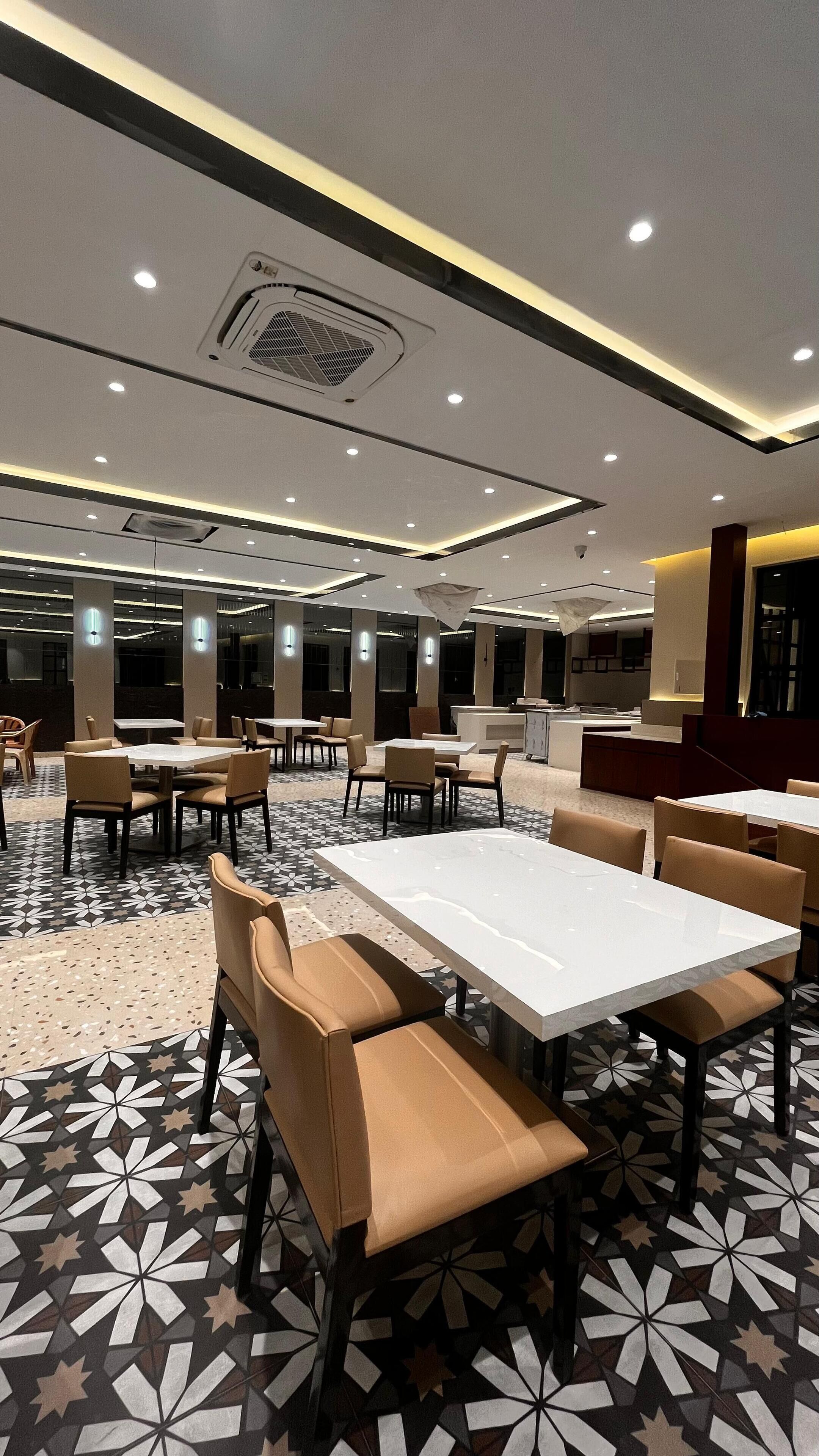 A spacious dining area with multiple tables and chairs, a patterned floor, and a modern ceiling design with recessed lighting.
