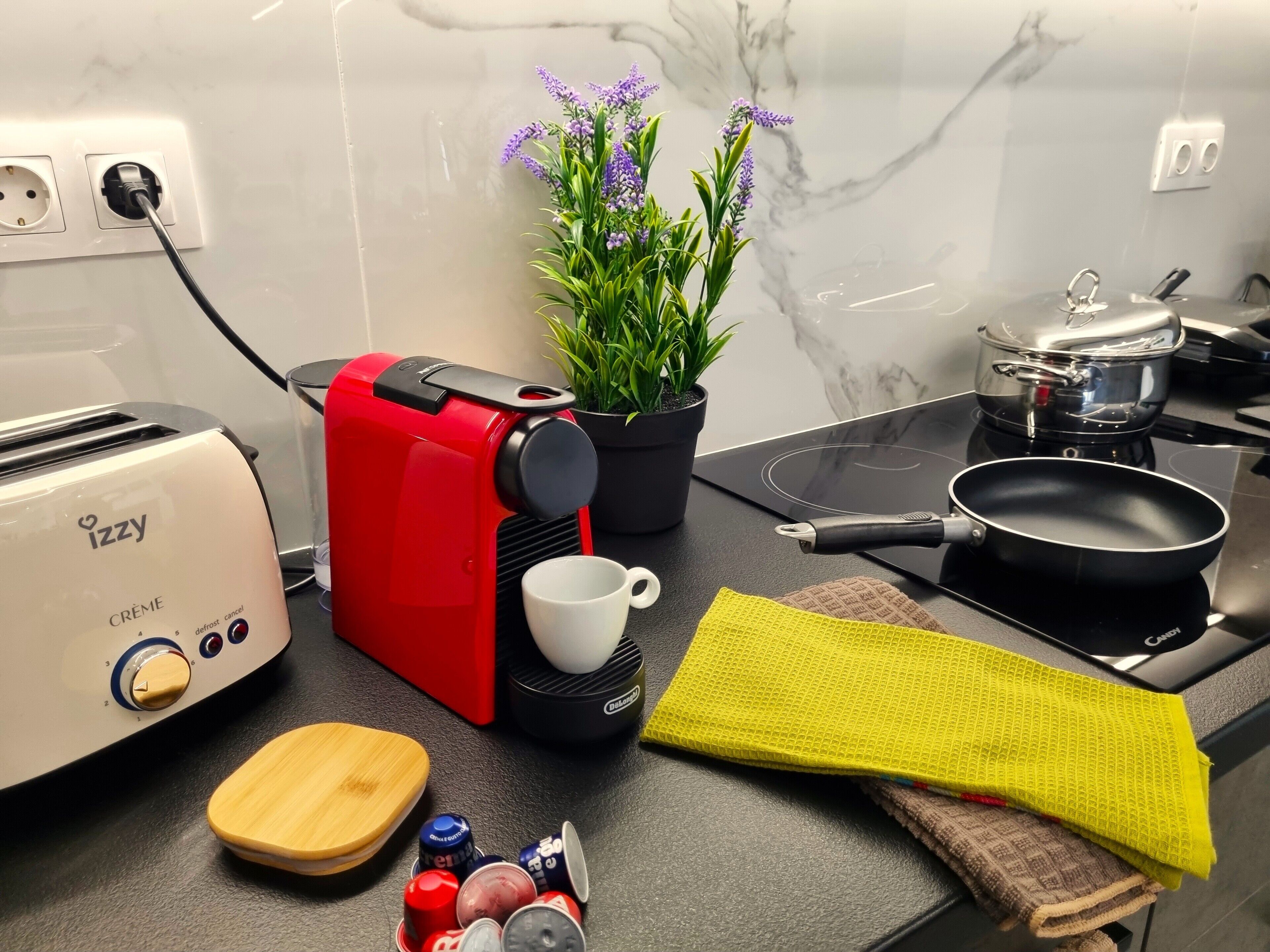 A kitchen with a toaster, a coffee maker, a frying pan, a pot, a potted plant, and a wooden cutting board.