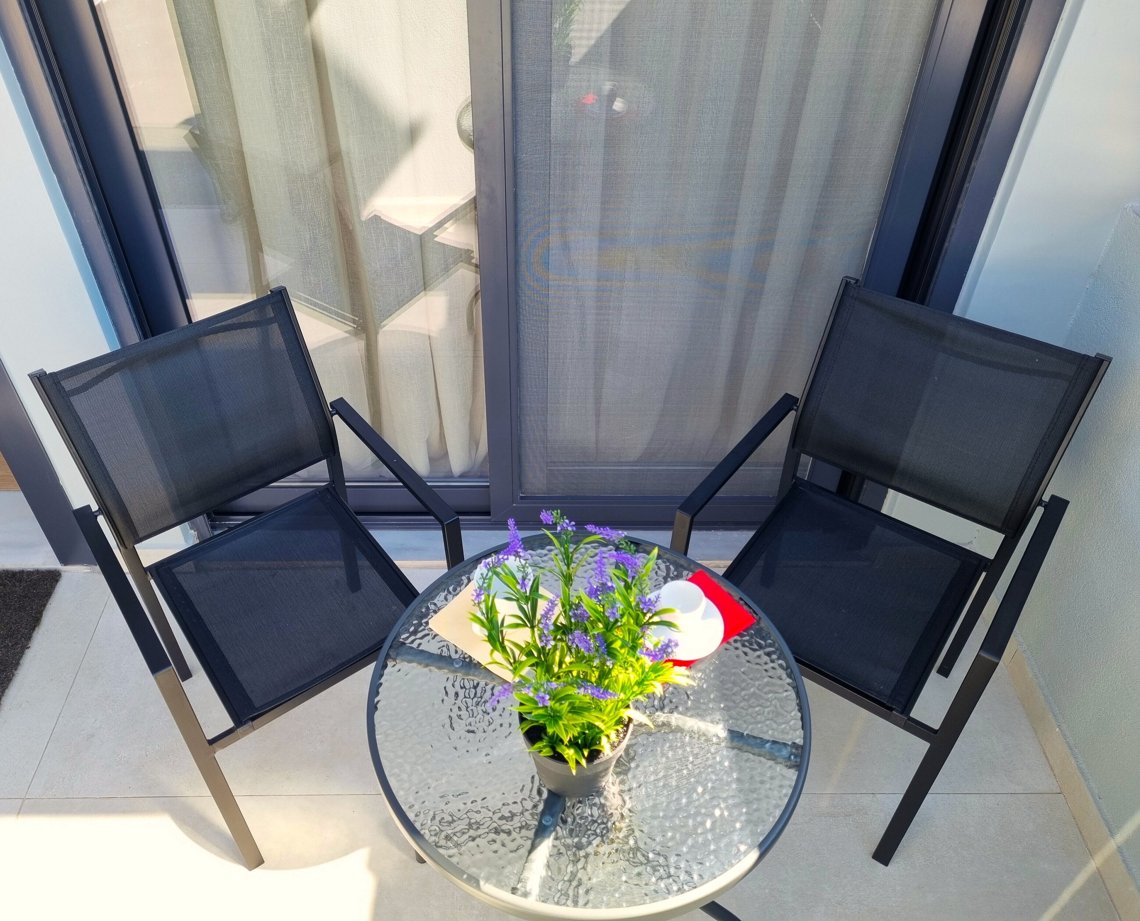 A glass table with two black chairs, a potted plant with purple flowers, and a red object on the table.