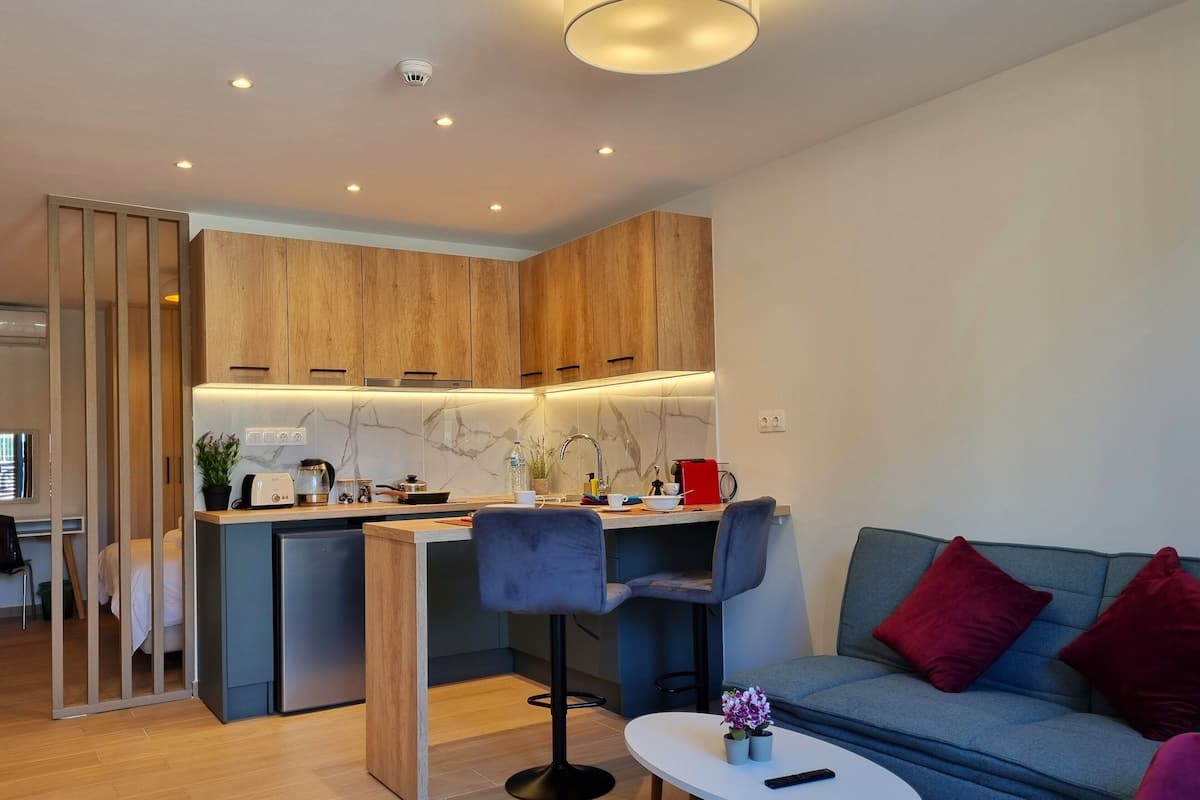 A modern kitchen with wooden cabinets, a marble backsplash, and recessed lighting. There is a dining area with a grey sofa and red cushions, and a small white table with a remote control.