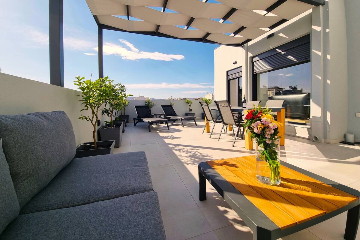 A rooftop terrace with a sofa, chairs, a table with a vase of flowers, and a grill.