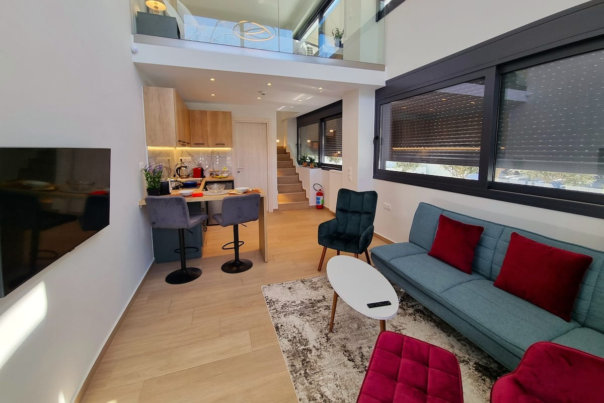 A modern apartment with a kitchen, dining area, and living room. It features a staircase connecting two floors, a large window with blinds, and a small balcony.